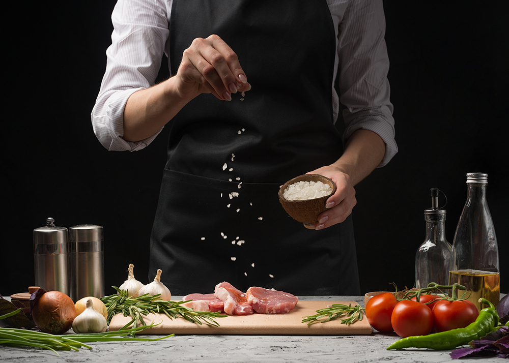 Chef salts steak grill pan. Preparing fresh beef or pork. Horizontal photo with a dark black background